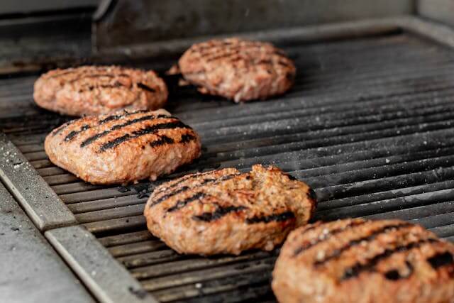 Five thick burger patties with grill marks cooking on a clean barbecue grill, illustrating proper grilling technique and even heat.