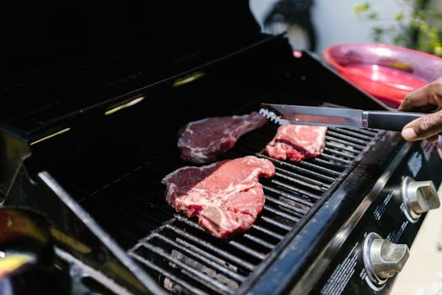 Person using tongs to grill three steaks on a gas barbecue, showing proper safe grilling technique and food handling.