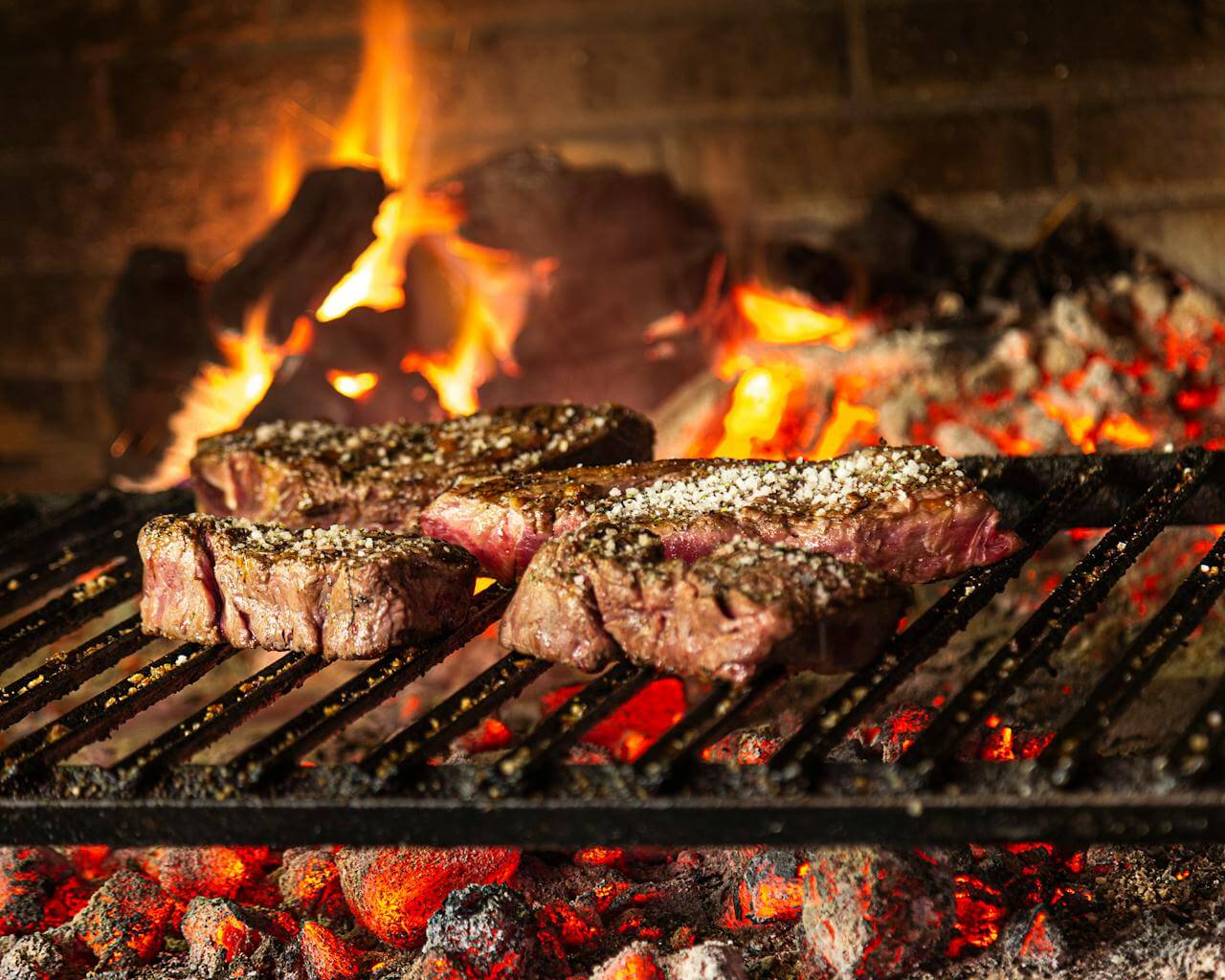 Three seasoned steaks cooking on a grill over hot coals and a wood fire, showing proper grilling technique and high heat for barbecue.